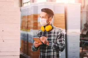 Portrait of man supervisor in medical face mask and protective headphones checking wood material inventory at storage. Young warehouse worker inspecting, counting woodwork stock. COVID-19 quarantine